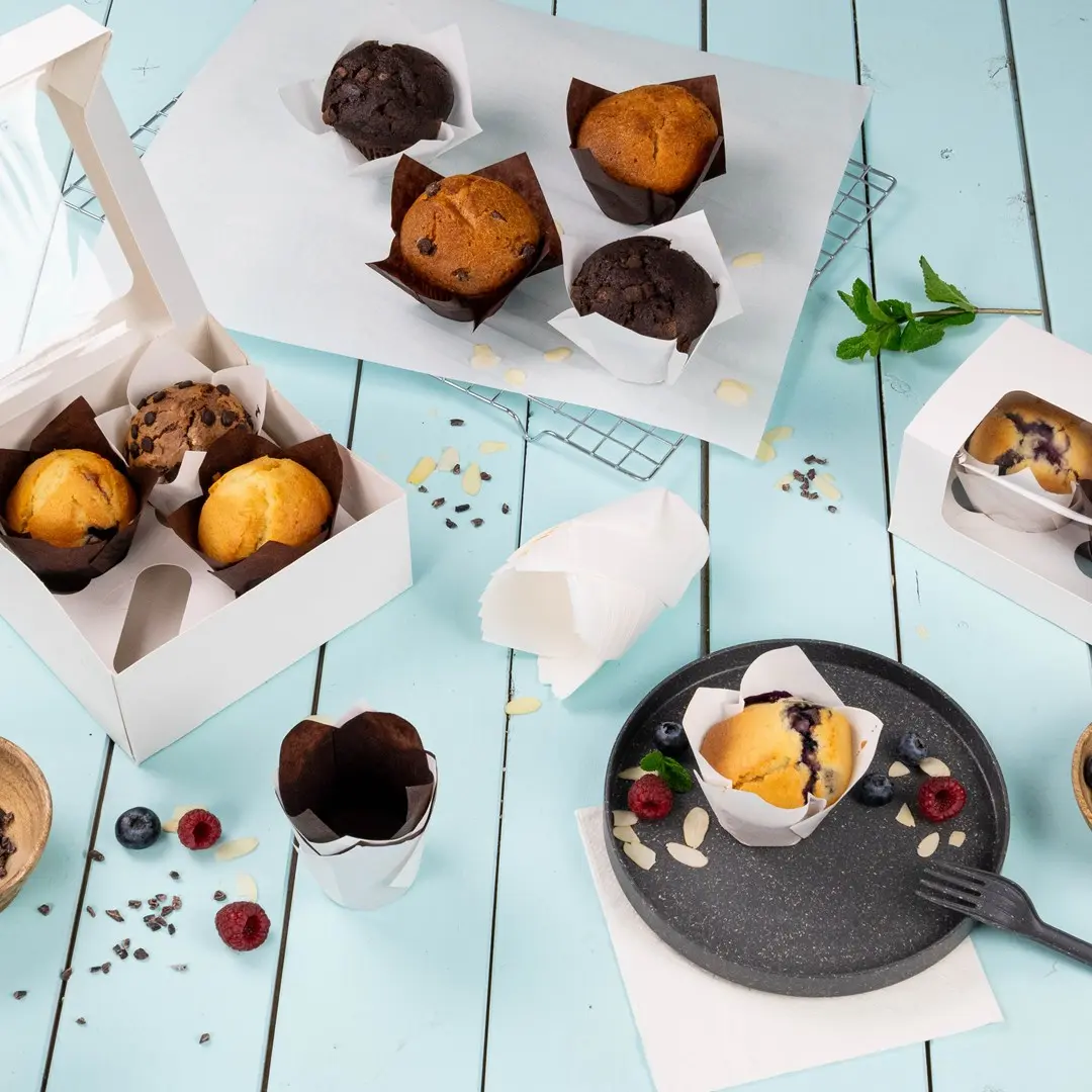 Muffin assortiti in involucri di carta su una superficie azzurra, con una scatola bianca e un piatto di muffin e bacche.