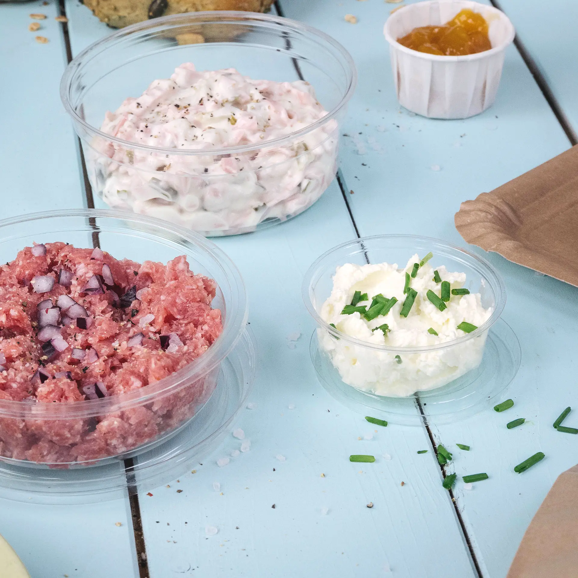 Tre bicchieri trasparenti con diversi alimenti su una superficie in legno azzurro chiaro. I bicchieri contengono carne macinata, una salsa bianca con erba cipollina e una salsa in un piccolo bicchiere.