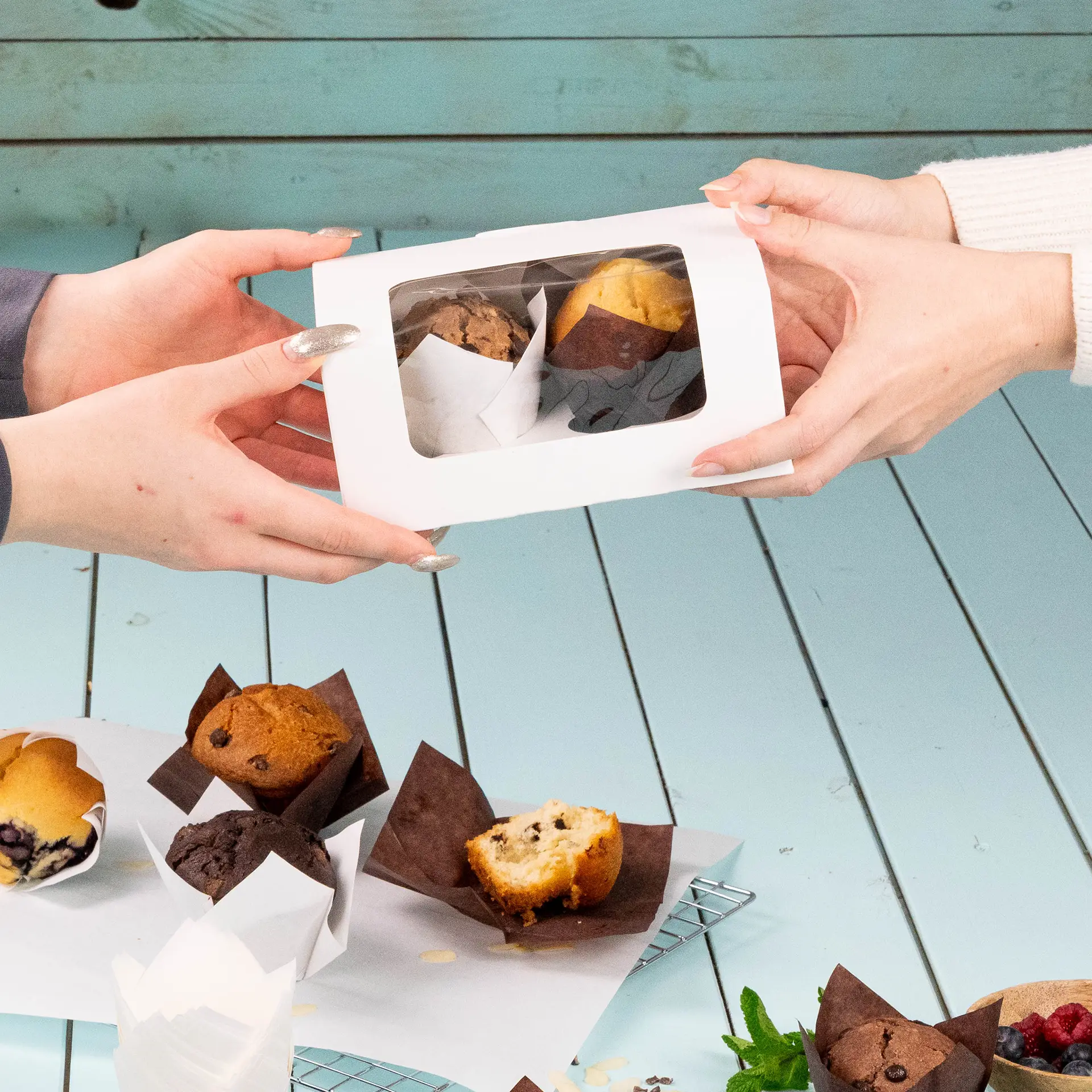 Scatola per cupcake bianca a due pezzi con finestra in cellulosa, consegnata con due muffin, su un tavolo azzurro.