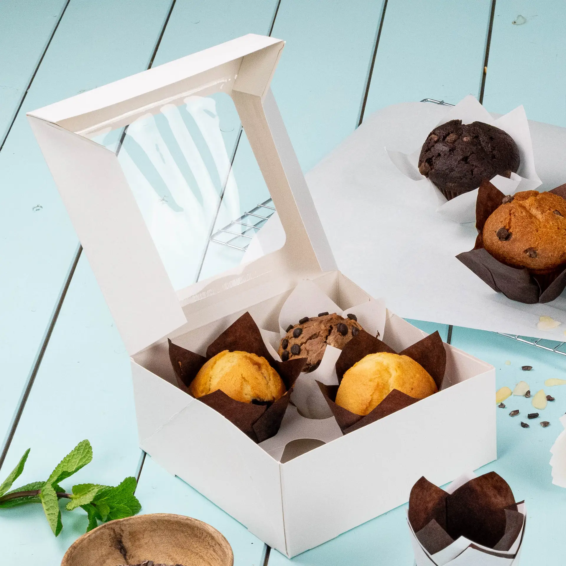 Scatola per cupcake bianca da 4 pezzi con una finestra in cellulosa, contenente muffin e un muffin al cioccolato, su una superficie blu chiaro.